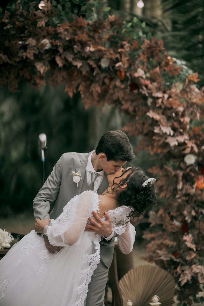 A couple shares a passionate kiss beneath a lush floral arch at their outdoor wedding ceremony.